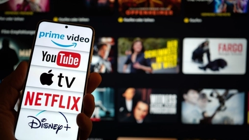 Close-up of a hand holding a smartphone with streaming platform logos on screen in front of a television